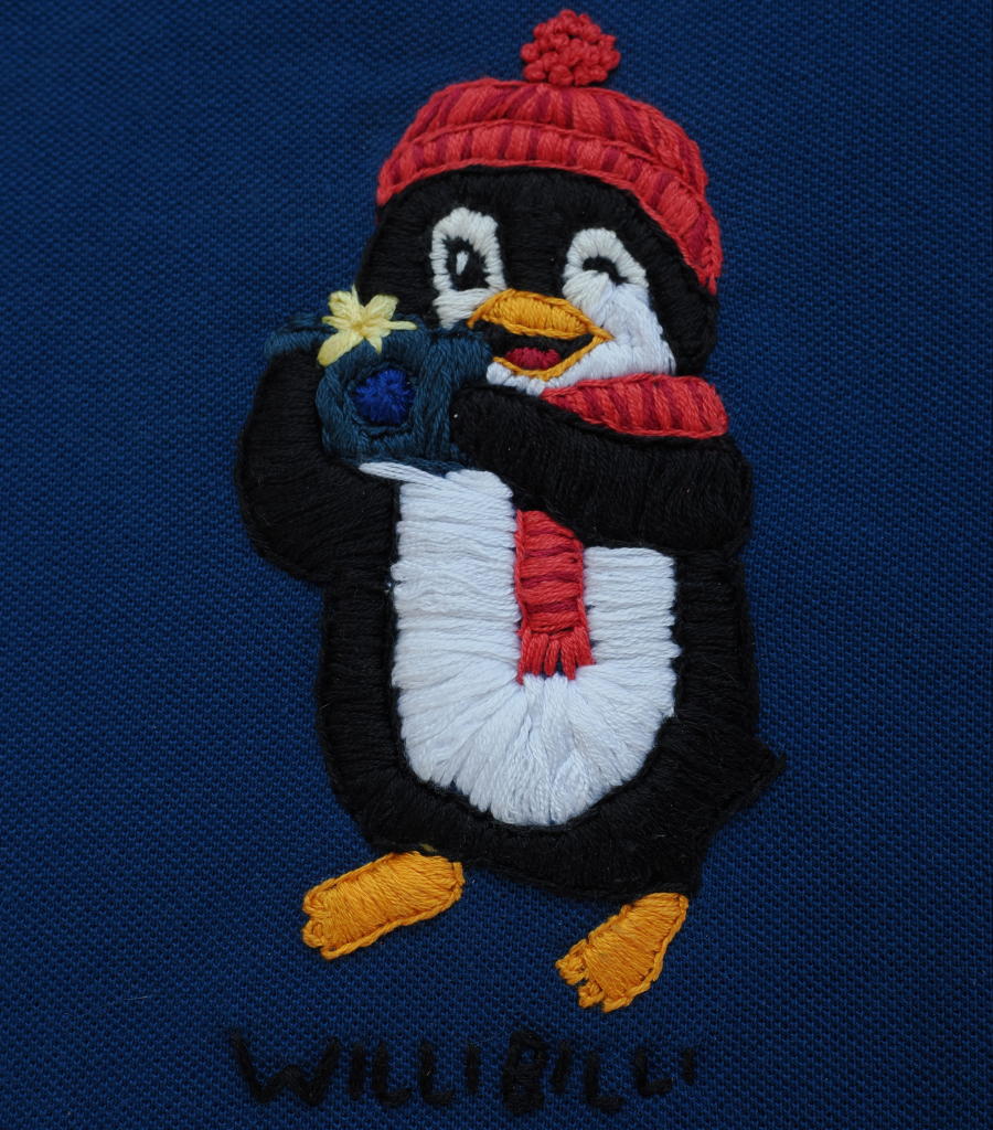 Auf dunkelblauem Hintergrund ist ein Pinguin mit roter Wollmütze, rotem Schal und einem Fotoapparat der gerade blitzt aufgestickt. Darunter ist in schwarzer Schrift WilliBilli aufgestickt.